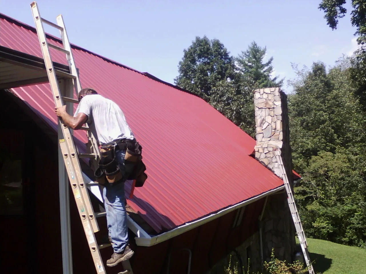 Roofer on a ladder beside a red metal roof and stone chimney — Soffit & Fascia Repair by Roofing Brothers in Jetson